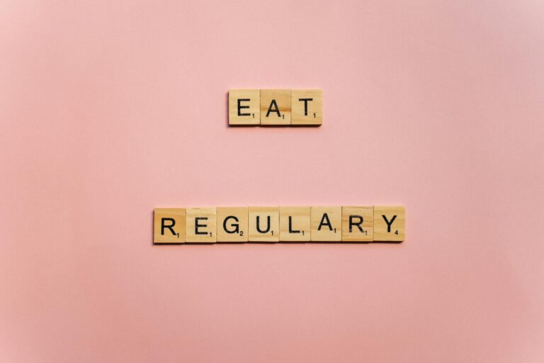 Wooden tiles spelling 'Eat Regularly' on a pink background, promoting healthy eating habits.